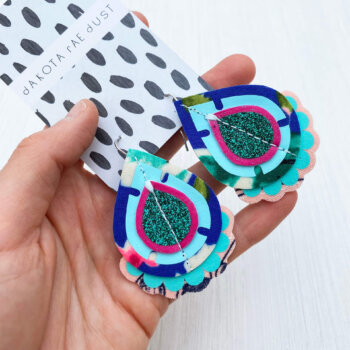 A pair of peacock blue, green and raspberry recycled fabric earrings held in a open hand