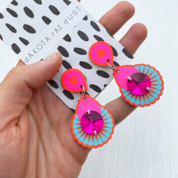 A pair of neon pink orange and blue mini jewel earrings with hot pink gems mounted on a black and white patterned, branded dakota rae dust card and held in a white woman's hand