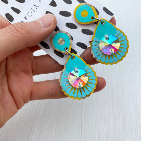 A pair of turquoise blue and yellow mini jewel earrings with iridescent gems mounted on a black and white patterned, branded dakota rae dust card and held in a white woman's hand