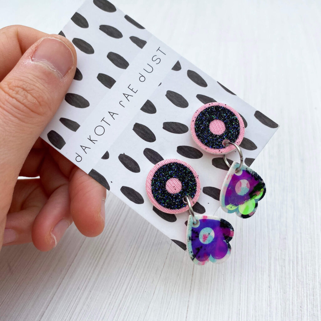 A close up of pair of pale pink, glittery black and iridescent small dangly studs mounted on a black and white patterned dakota rae dust branded card held in an open hand