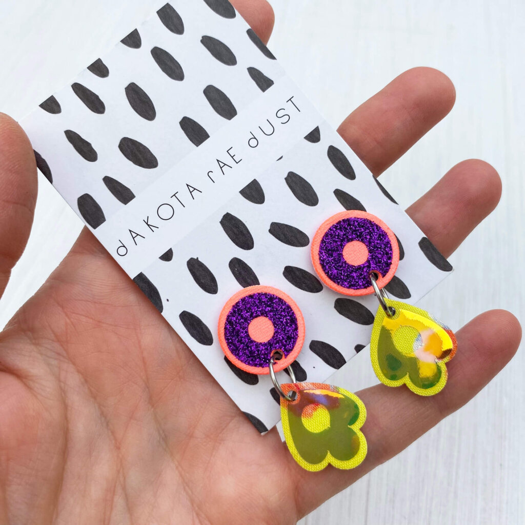 A close up of pair of small dangly stud earrings in tropical peach, purple and yellow mounted on a black and white patterned dakota rae dust branded card held in an open hand