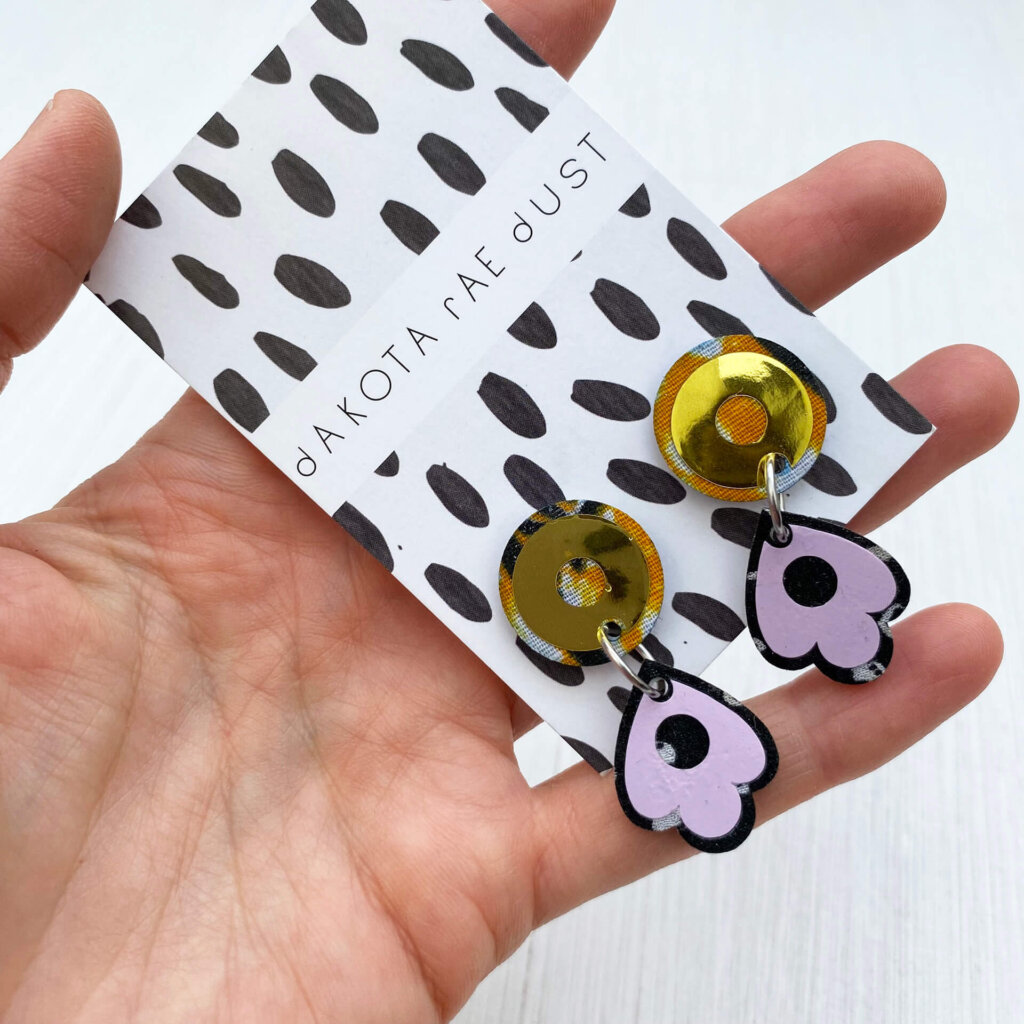 A close up of pair of gold and lilac small dangly studs mounted on a black and white patterned dakota rae dust branded card held in an open hand