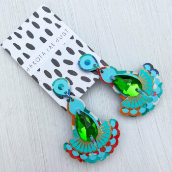 A pair of turquoise green jewel earrings mounted on a black and white patterned dakota rae dust branded card are lying on an off white background