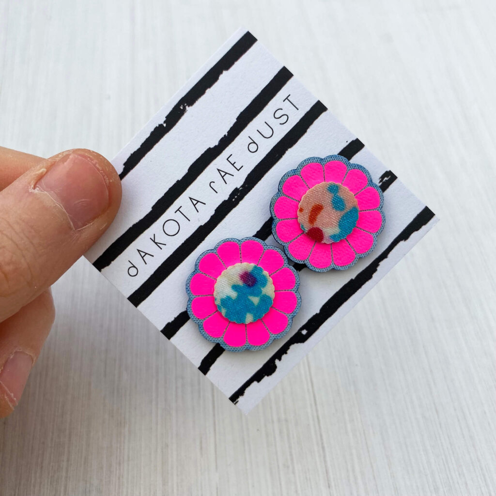 A pair of fluoro pink floral studs mounted on a black and white patterned, dakota rae dust branded card are held by a just visible thumb and forefinger