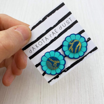 A pair of beautiful blue flower studs mounted on a black and white patterned, dakota rae dust branded card are held by a just visible thumb and forefinger
