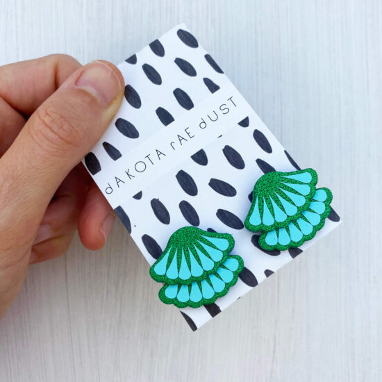 a pair of turquoise green tiered frill studs, mounted on a black and white patterned, dakota rae dust branded card and held against an off white background between a white thumb and forefinger