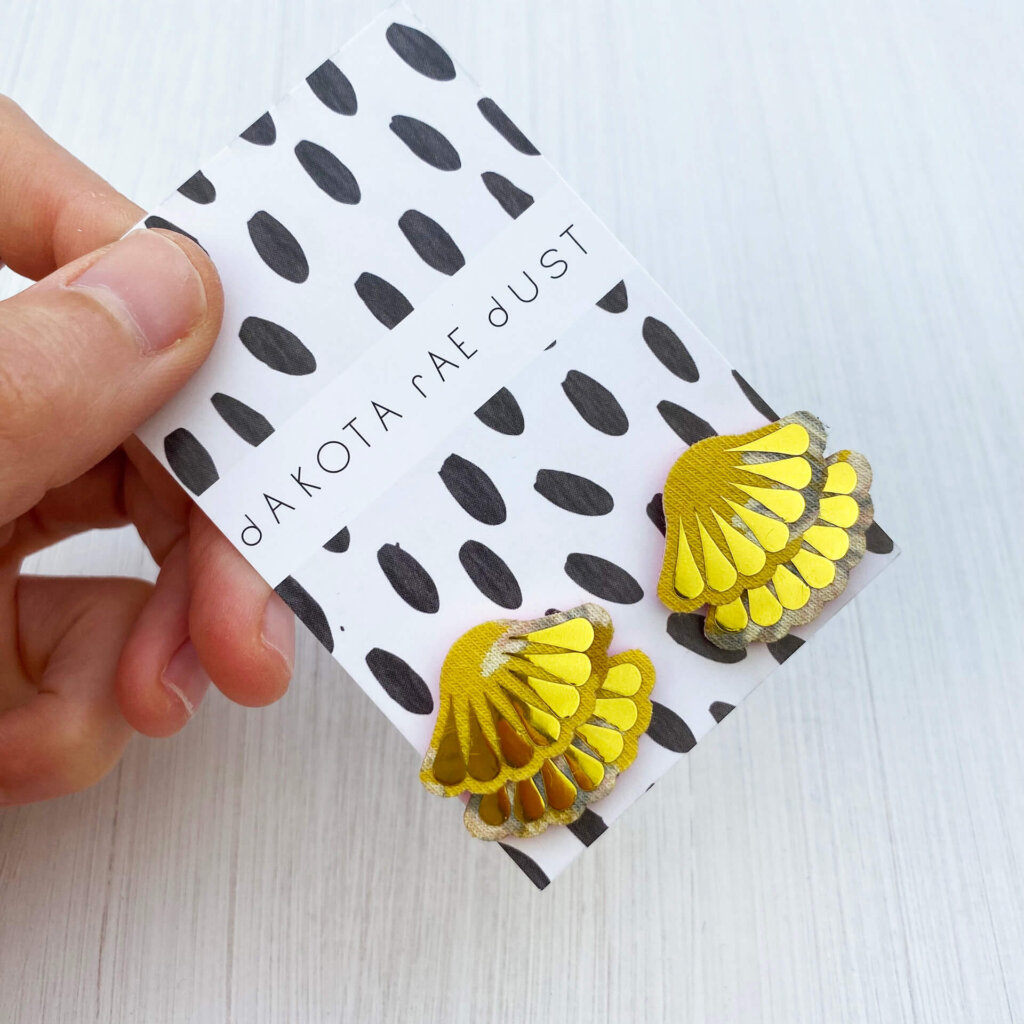 a pair of gold and yellow tiered frill studs, mounted on a black and white patterned, dakota rae dust branded card and held against an off white background between a white thumb and forefinger