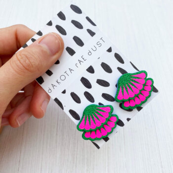 a pair of fluorescent pink and green tiered frill studs, mounted on a black and white patterned, dakota rae dust branded card and held against an off white background between a white thumb and forefinger