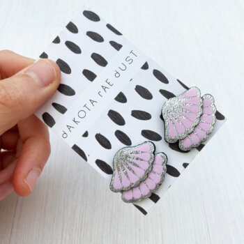 a pair of silver and pastel pink tiered frill studs, mounted on a black and white patterned, dakota rae dust branded card and held against an off white background between a white thumb and forefinger