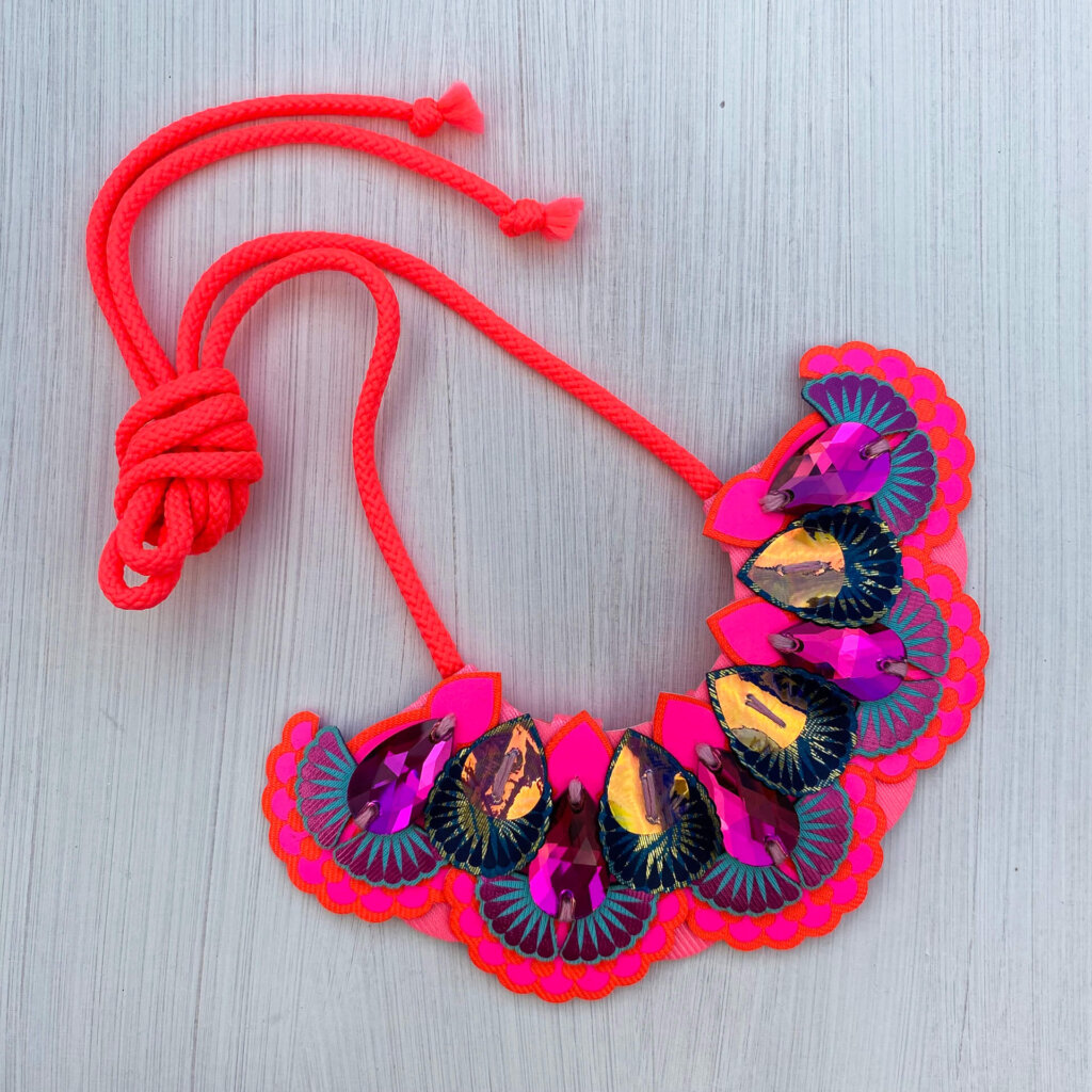 A neon pink, orange and navy oversize statement bib necklace with neon coral cord ties and dark iridescent jewels displayed an off white background