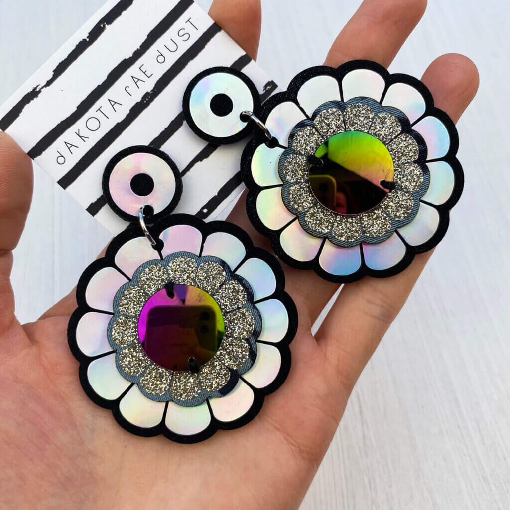 A pair of oversize holographic flower earrings with petrol coloured discs in the centre, mounted on a stripey dakota rae dust branded card are held in an open hand.