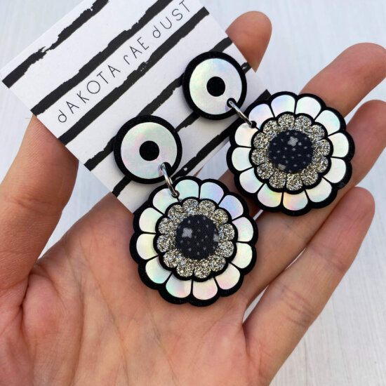 A pair of mini holographic flower earrings mounted on a black and white stripey dakota rae dust branded card are held in an open hand