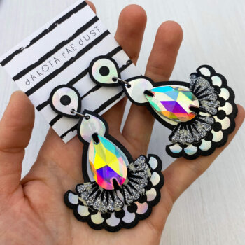 A pair of holographic iridescent jewel earrings mounted on a dakota rae dust branded card are held in an open hand
