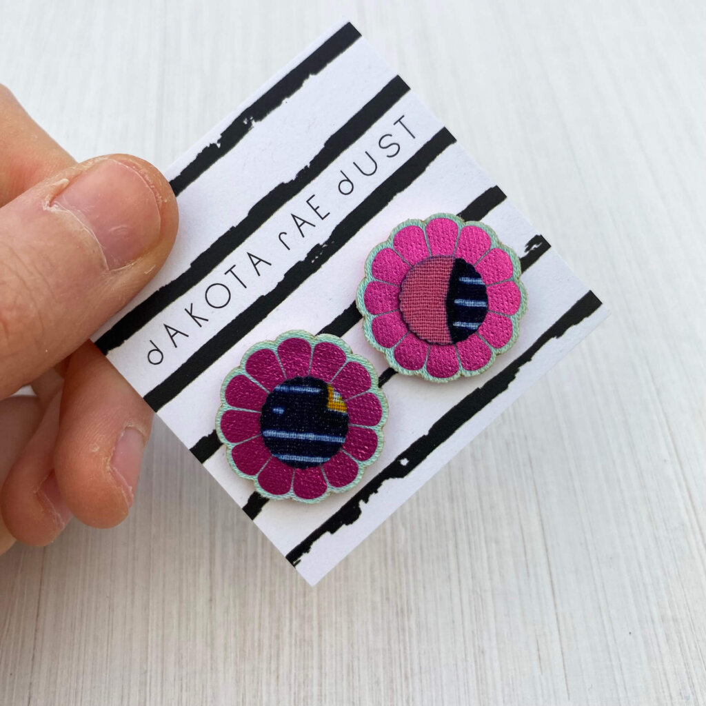 A pair of metallic pink flower studs mounted on a black and white stripey, dakota rae dust branded card are held by a just visible thumb and forefinger