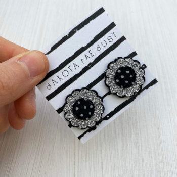 A pair of glittery silver flower studs mounted on a dakota rae dust branded card are held against an off white background in a just visible thumb and forefinger.