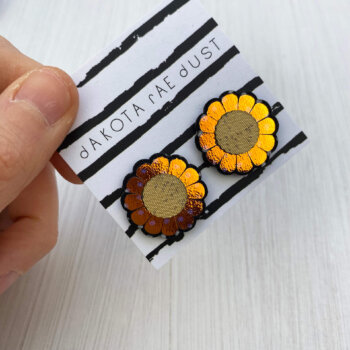 A pair of iridescent gold flower studs mounted on a black and white, dakota rae dust branded card are held between a just visible thumb and forefinger