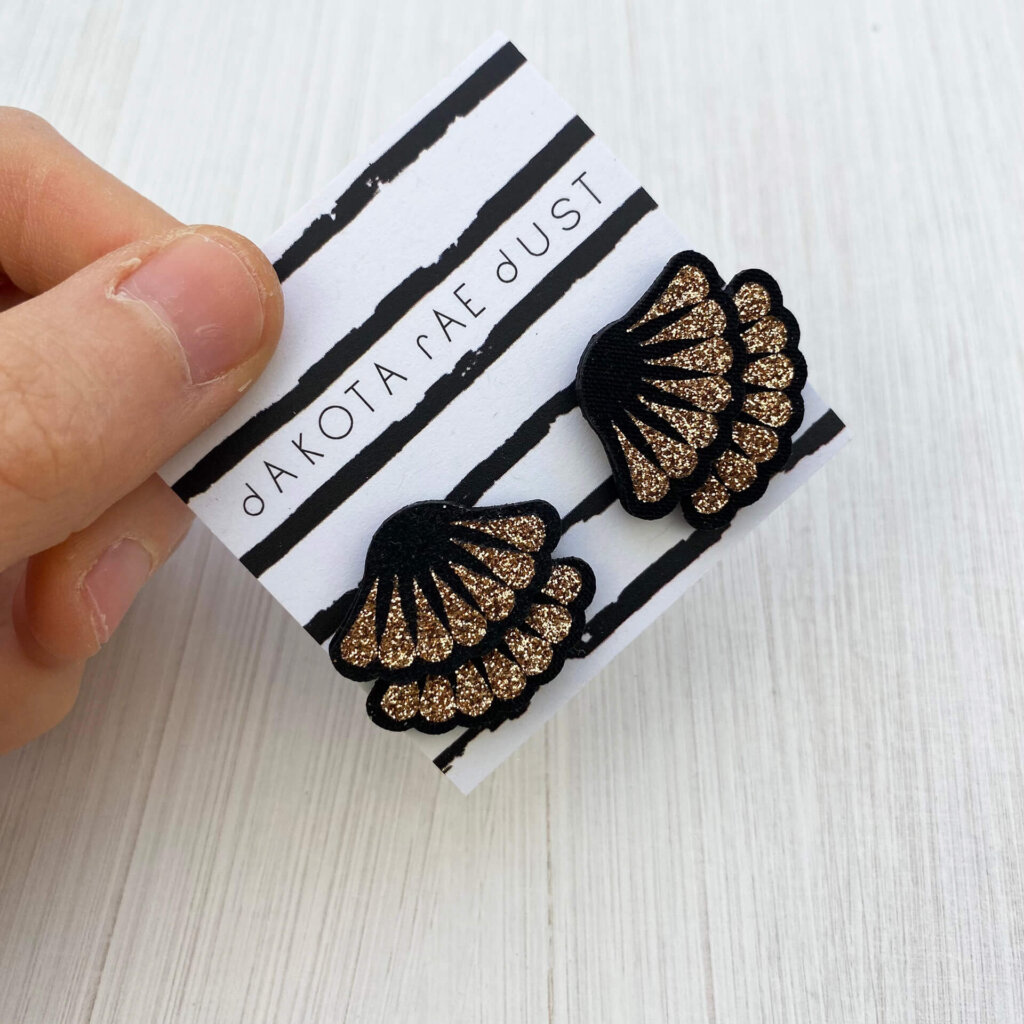 A pair of antique gold glitter frill studs mounted on a dakota rae dust branded card are held by a just visible thumb and forefinger.
