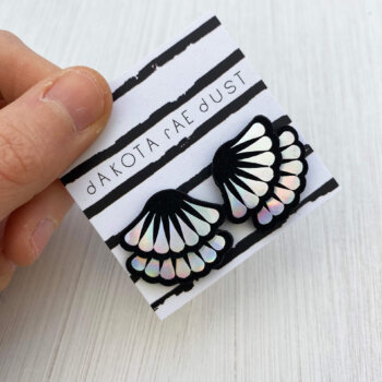 A pair of holographic frill studs mounted on a dakota rae dust branded card are held by a just visible thumb and forefinger.