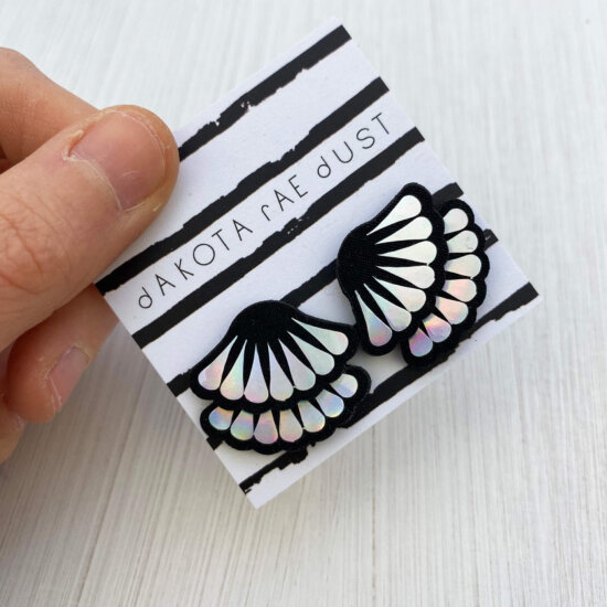 A pair of holographic frill studs mounted on a dakota rae dust branded card are held by a just visible thumb and forefinger.