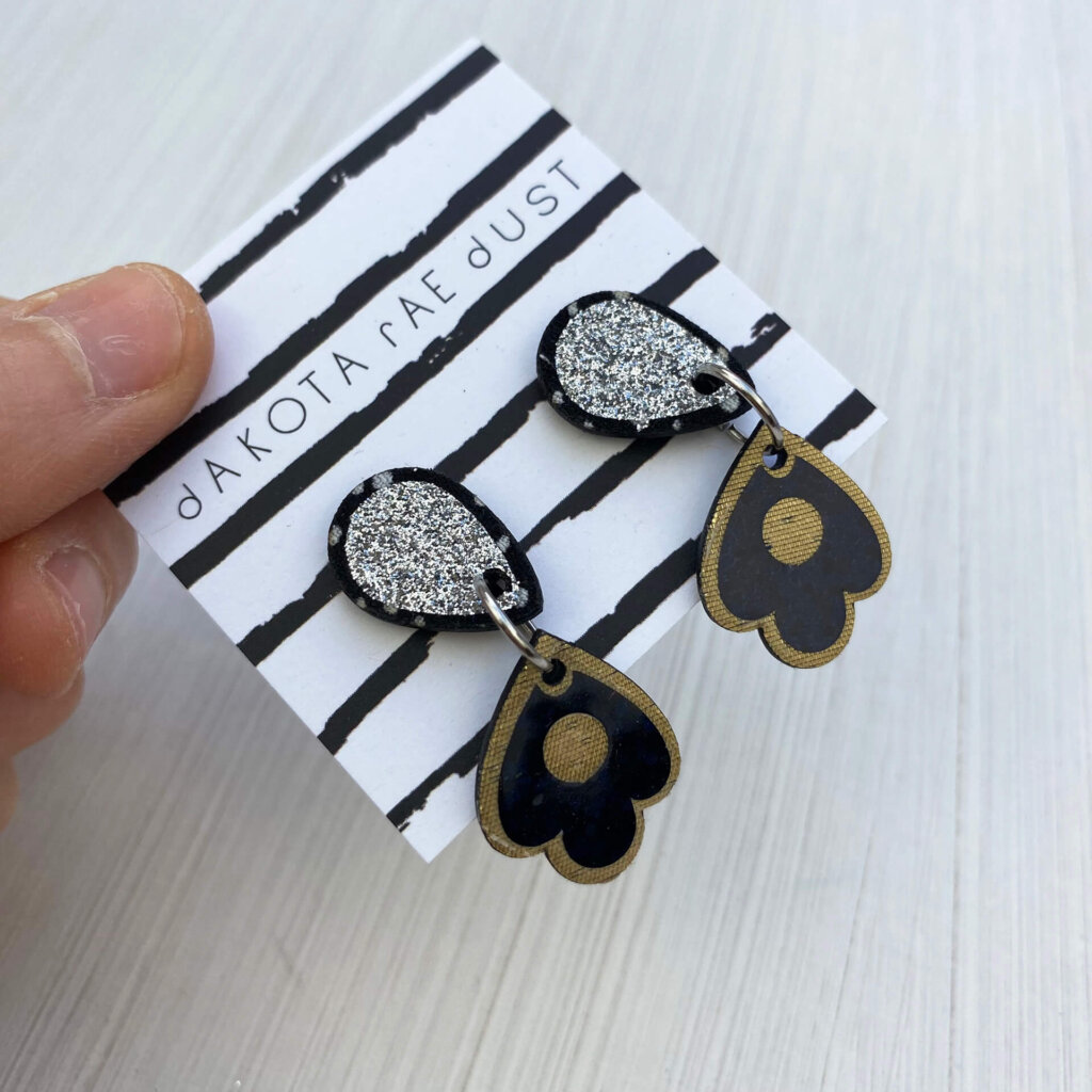 A pair of gold, black and silver sparkly dangly studs mounted on a dakota rae dust branded card are held out for the camera between a just visible thumb and forefinger