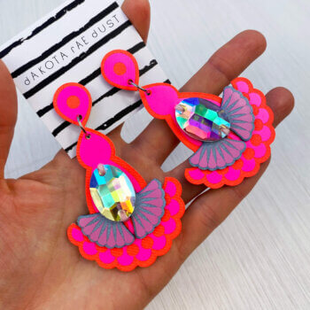A pair of iridescent jewel earrings in neon pink and orange are held in an open hand
