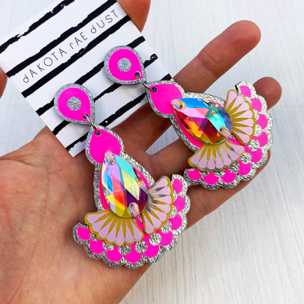 A pair of iridescent, neon pink and silver jewel earrings are held in an open hand