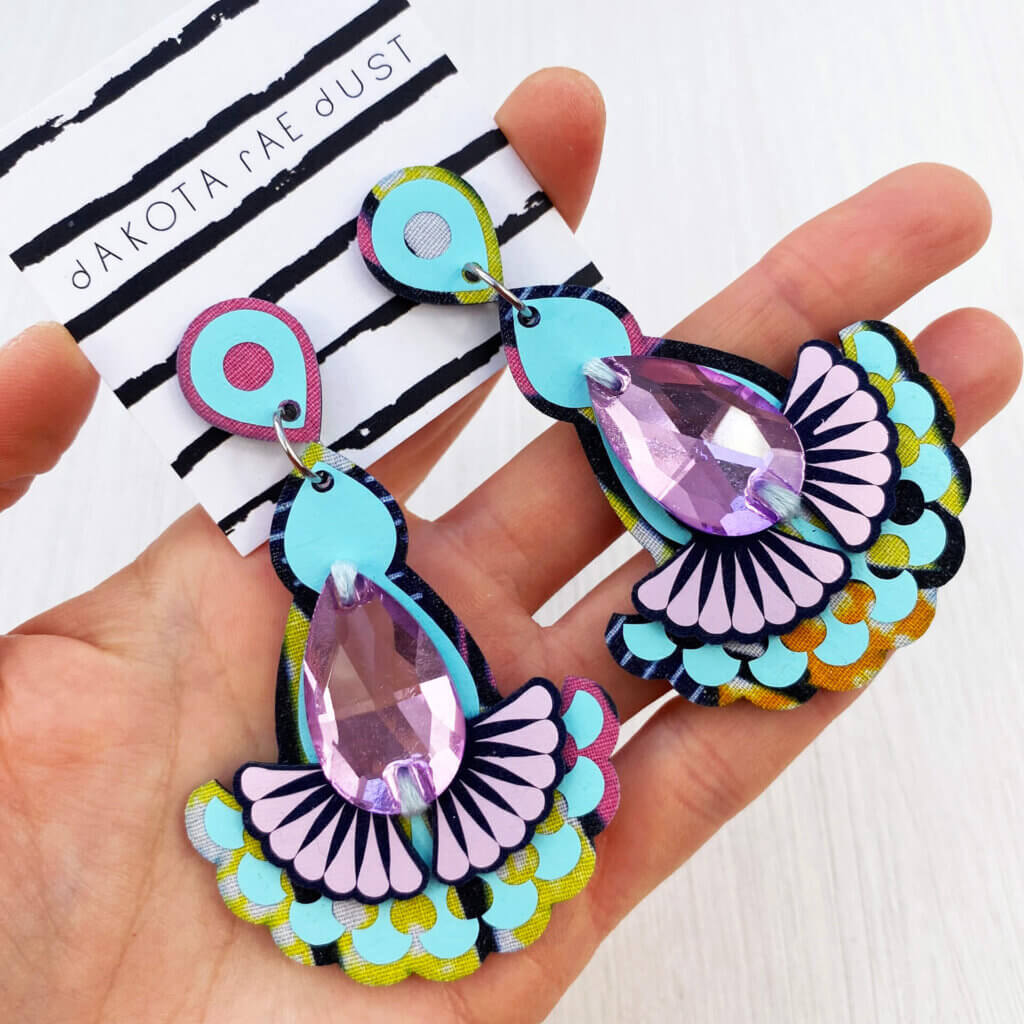 A pair of light blue and lilac jewel earrings are held in an open hand
