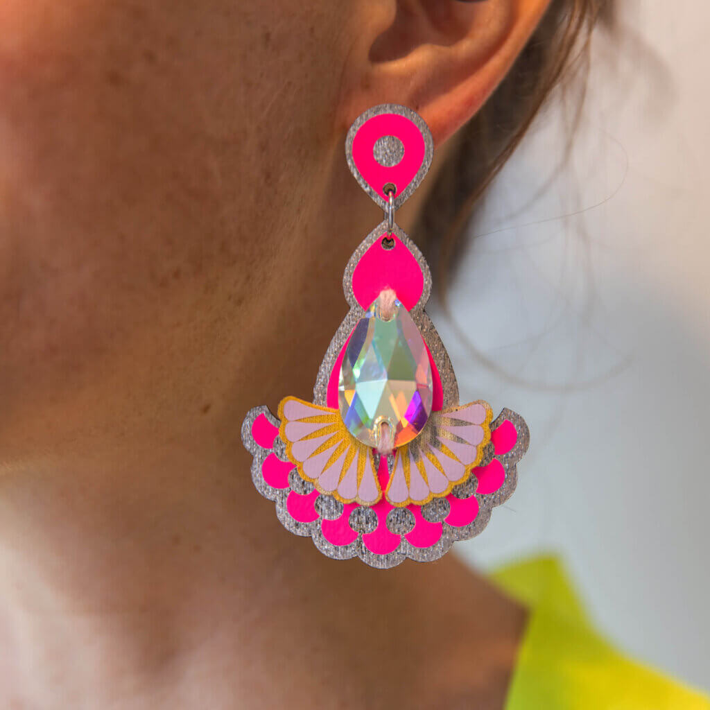 a close up of a neon pink and silver statement jewel earrings with an iridescent jewel hanging in a woman's ear.