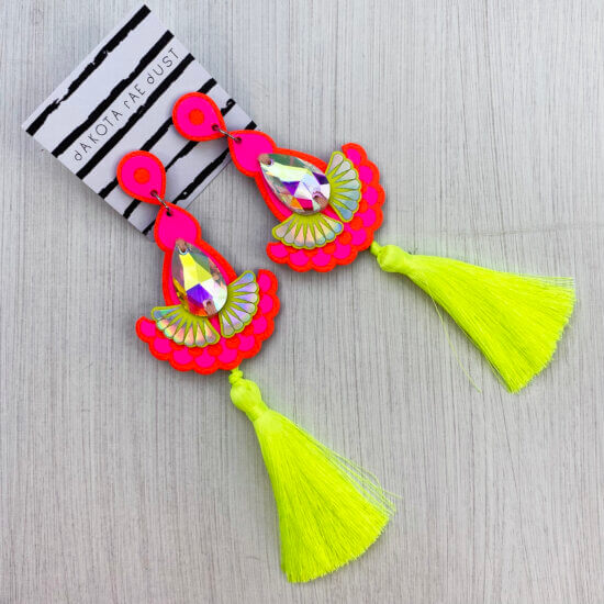 A pair of bright orange, pink and fluorescent yellow jewel and tassel earrings are lying on an off white background