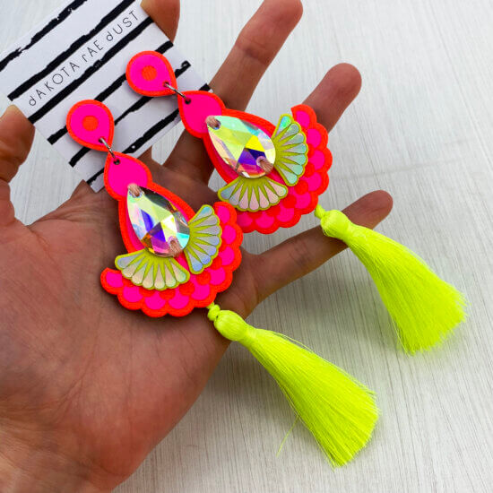 A pair of orange, pink and yellow fluorescent tassel earrings are held in an open hand
