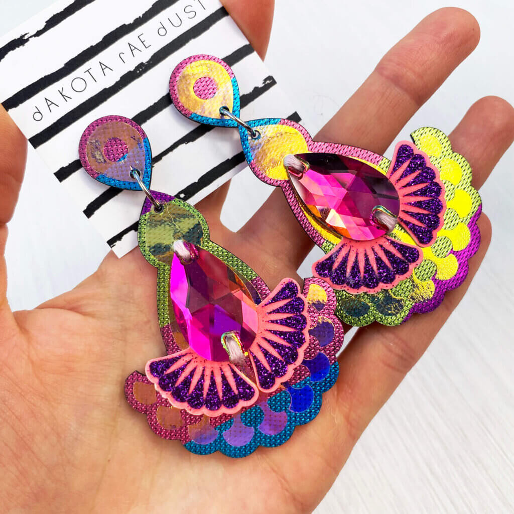 A pair of pink, turquoise, lime green and purple Tropical iridescent jewel earrings are held in an open hand.