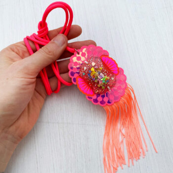A pink and peach custom cast giant jewel pendant necklace with a neon coral fringe lying on an off white background