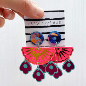 A colourful pair of chandelier style, jangly droplet earrings mounted on a black and white striped, dakota rae dust branded card are held between a just visible thumb and forefinger