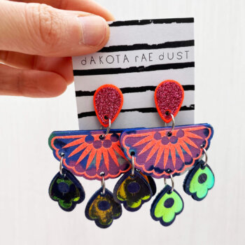 A pair of colourful chandelier style, jangly droplet earrings mounted on a black and white striped, dakota rae dust branded card are held between a just visible thumb and forefinger