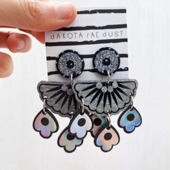 A pair of chandelier style, jangly droplet earrings mounted on a black and white striped, dakota rae dust branded card are held between a just visible thumb and forefinger