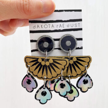 A pair of chandelier style, jangly droplet earrings mounted on a black and white striped, dakota rae dust branded card are held between a just visible thumb and forefinger