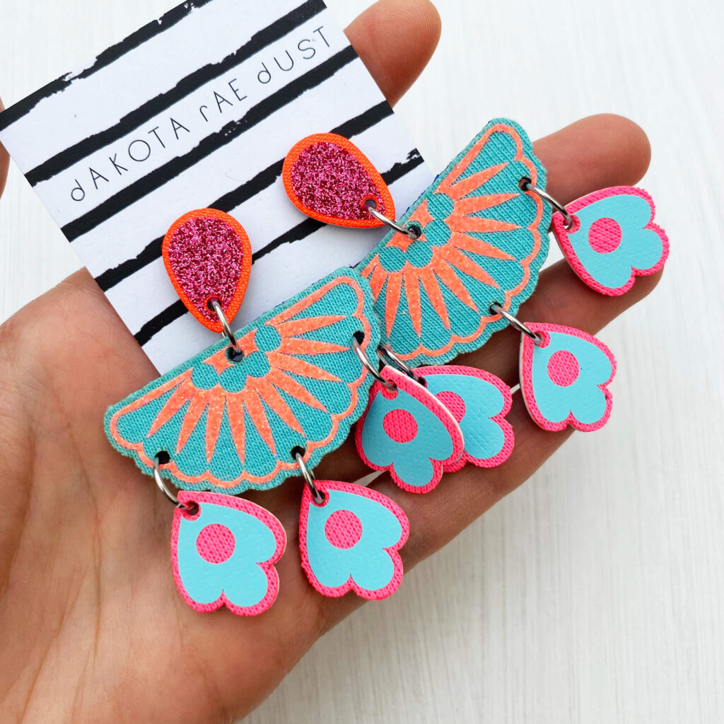 A pair of turquoise, pink and orange chandelier style statement earrings are held in an open hand