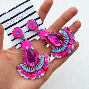 A pair of fluoro pink and floral jewel earrings are held in an open hand