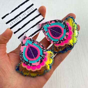 A pair of oversize secondhand fabric earrings mounted on a black and white patterned, dakota rae dust branded card are held in an open hand