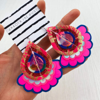 A pair of pink blue gold oversize earrings mounted on a black and white patterned, dakota rae dust branded card are held in an open hand