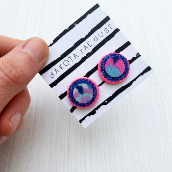 a pair of neon pink and glittery blue small round studs mounted on a black and white striped, dakota rae dust branded card are held between a just visible thumb and forefinger