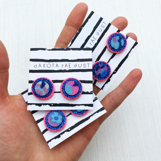 Three pairs of pink and blue small circular studs mounted on black and white striped, dakota rae dust branded cards and held in an open hand.