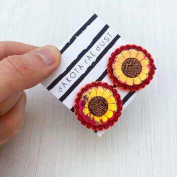 A pair of oversize red gold studs mounted on a dakota rae dust branded card are held between a just visible thumb and forefinger