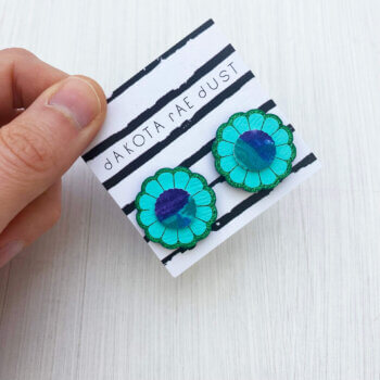 A pair of turquoise green flower studs mounted on a dakota rae dust branded card are held against an off white background in a just visible thumb and forefinger.
